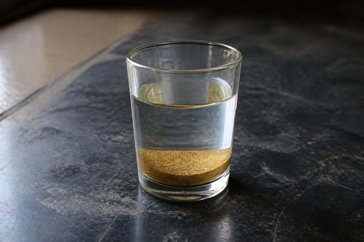 A glass of water with coriander powder settled into an even layer on the bottom and a clear surface