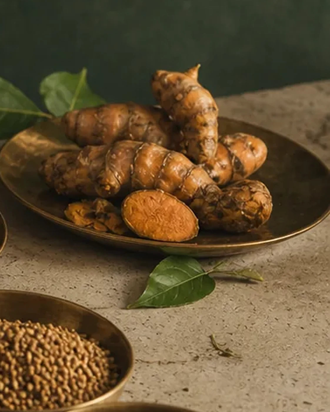 Whole turmeric roots on a brass plate beside a bowl of coriander seed